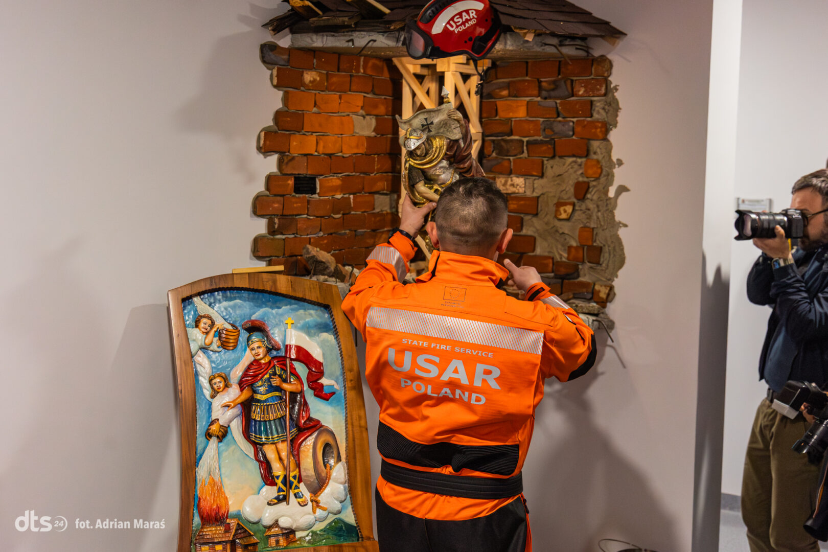USAR Poland firefighter in an orange uniform reattaching a statue atop rubble, as a photographer records the scene on the right.
