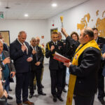Officials and uniformed personnel gather in a hallway as a man in a black suit with a yellow stole raises a brush for a blessing, holding a red folder nearby.
