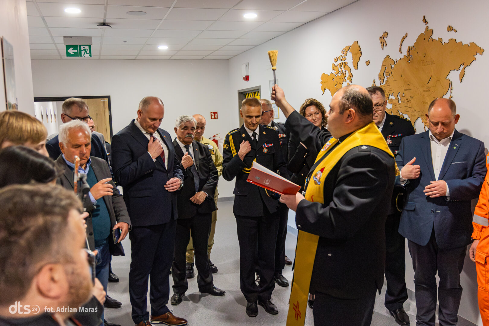 Officials and uniformed personnel gather in a hallway as a man in a black suit with a yellow stole raises a brush for a blessing, holding a red folder nearby.