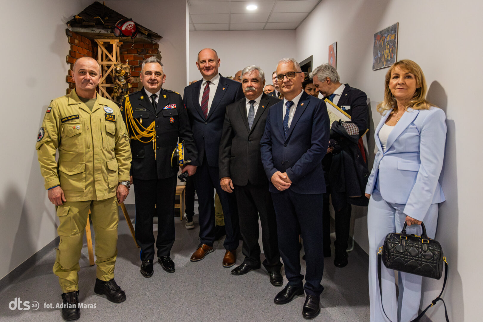 Group of uniformed and civilian officials posing in a hallway during a formal event, with a display structure in the background.