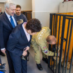 A group of officials in suits and a worker in a kennel lean over to pet a dog inside a metal enclosure at a shelter.
