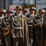 A group of soldiers in brown ceremonial uniforms with helmets, standing in formation with white cords and green badges. Centered figure holds a silver clarinet or woodwind instrument.
