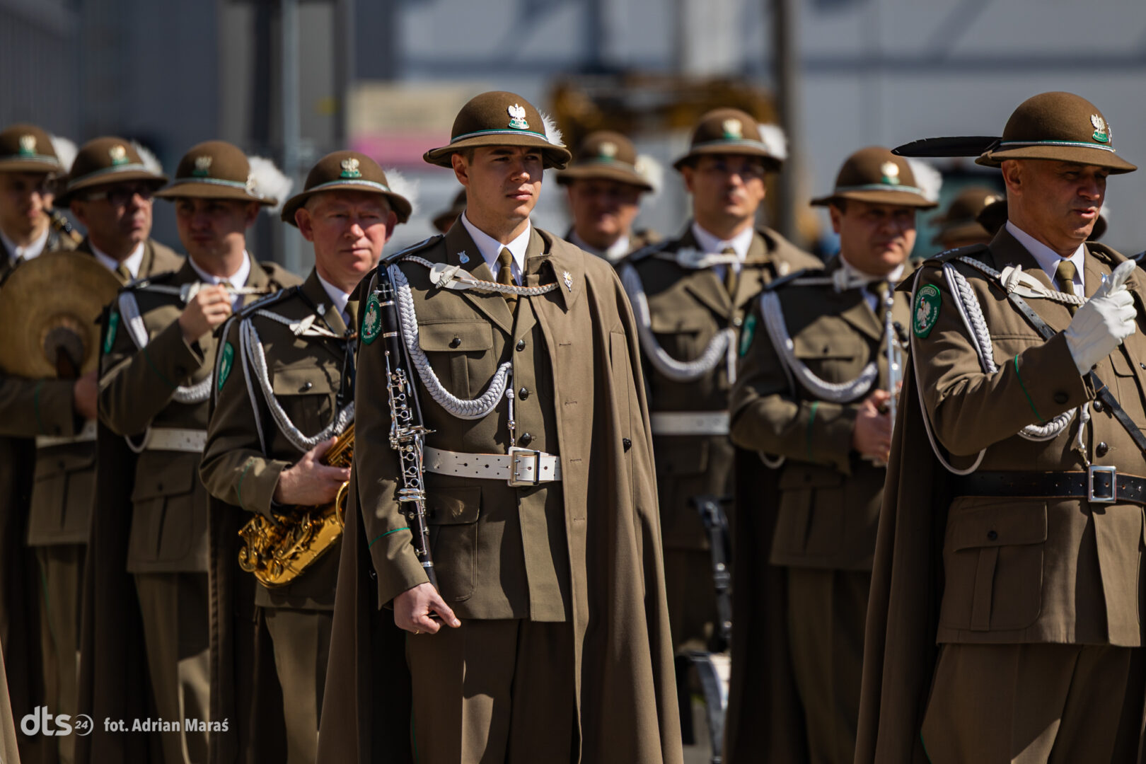 A group of soldiers in brown ceremonial uniforms with helmets, standing in formation with white cords and green badges. Centered figure holds a silver clarinet or woodwind instrument.