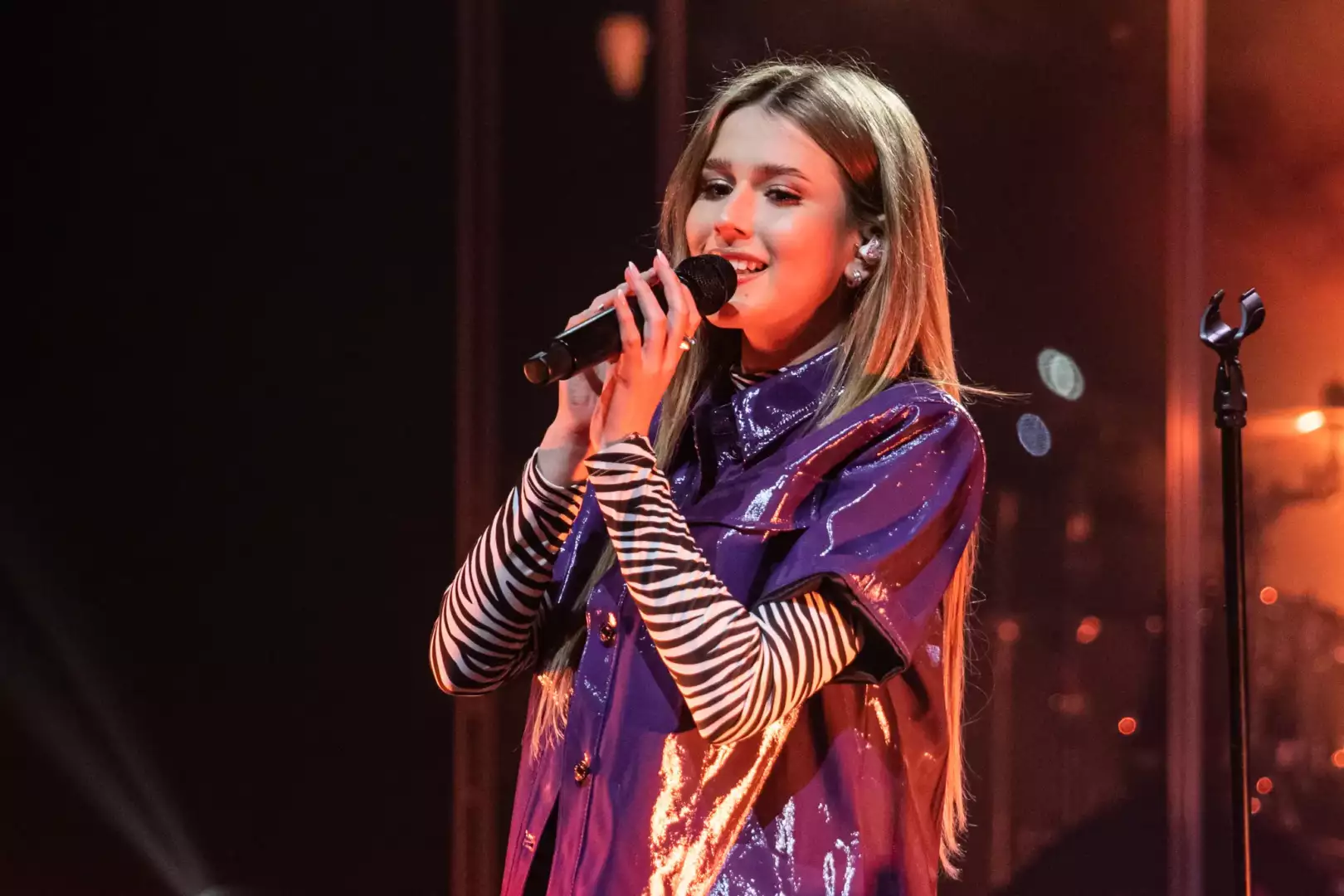 Female singer on stage, holding a microphone and performing in a shiny purple jacket with striped sleeves.