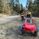 Rescue worker in a high‑visibility jacket walks beside a trailer carrying an ATV on a dirt road in a forested area with a fire truck nearby.