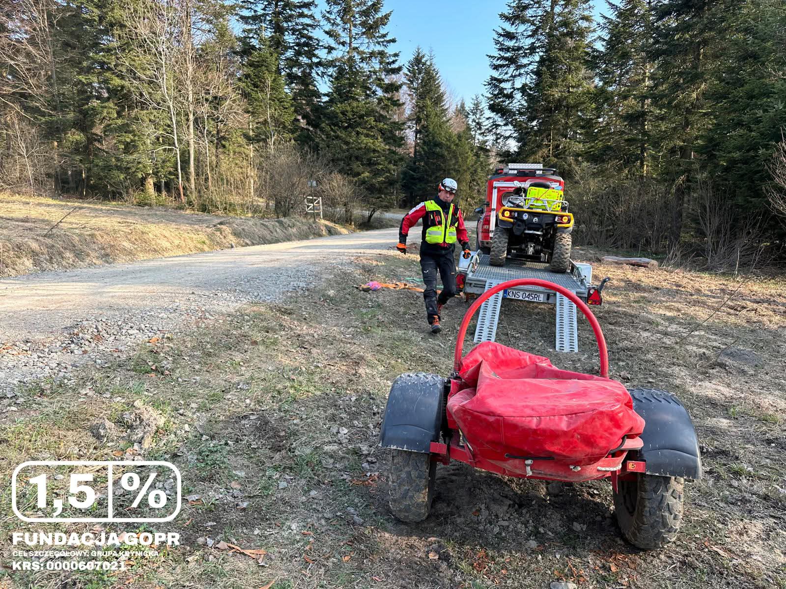 Rescue worker in a high‑visibility jacket walks beside a trailer carrying an ATV on a dirt road in a forested area with a fire truck nearby.