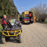 Emergency rescue scene: a yellow off-road quad bike in foreground, an ambulance with open doors and responders near a dirt road, forested area on left and bare trees on right, with Polish 1.5% fundraising graphic and GOPR foundation text in corner.
