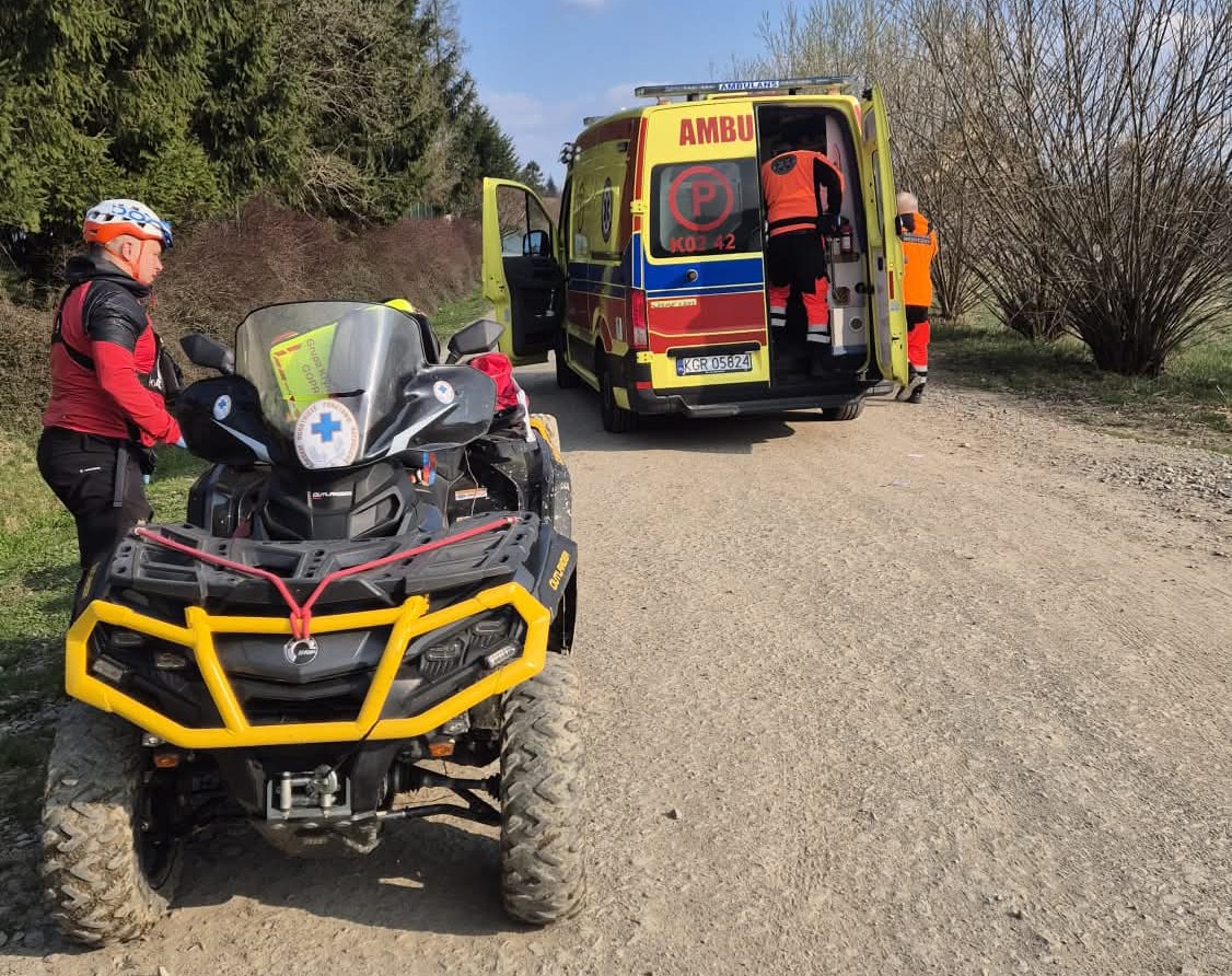 Emergency responders load a patient into a yellow ambulance on a dirt road, with a rugged ATV nearby.