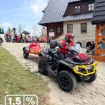 Man in a red jacket rides a black all-terrain vehicle with a red trailer along a stone path outside a rustic building; a crowd gathers in the background, and a white text overlay shows '1,5% FUNDACJA GOPR'.