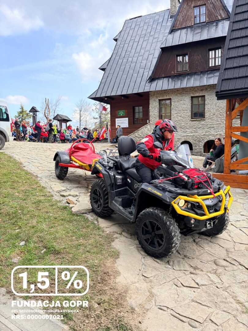 Man in a red jacket rides a black all-terrain vehicle with a red trailer along a stone path outside a rustic building; a crowd gathers in the background, and a white text overlay shows '1,5% FUNDACJA GOPR'.