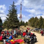 Crowd gathered at a forested hilltop festival, with a tall red-and-white communications tower and parked cars in the background.