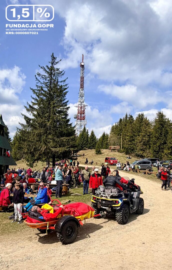 Crowd gathered at a forested hilltop festival, with a tall red-and-white communications tower and parked cars in the background.