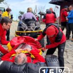 Emergency responders in red jackets load an injured person into a wheeled stretcher on a stone plaza, with onlookers and a wooden building nearby.