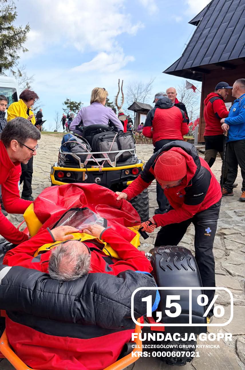 Emergency responders in red jackets load an injured person into a wheeled stretcher on a stone plaza, with onlookers and a wooden building nearby.