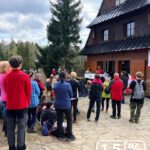 Outdoor gathering in front of a wooden mountain hut, people listening to a talk at a fundraiser.