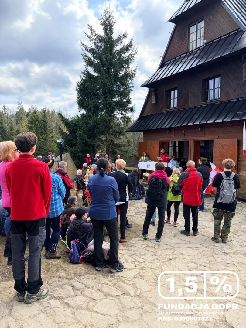Outdoor gathering in front of a wooden mountain hut, people listening to a talk at a fundraiser.
