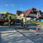Emergency responders and a yellow ambulance at a street crash scene with a toppled motorcycle and traffic cone on a crosswalk.