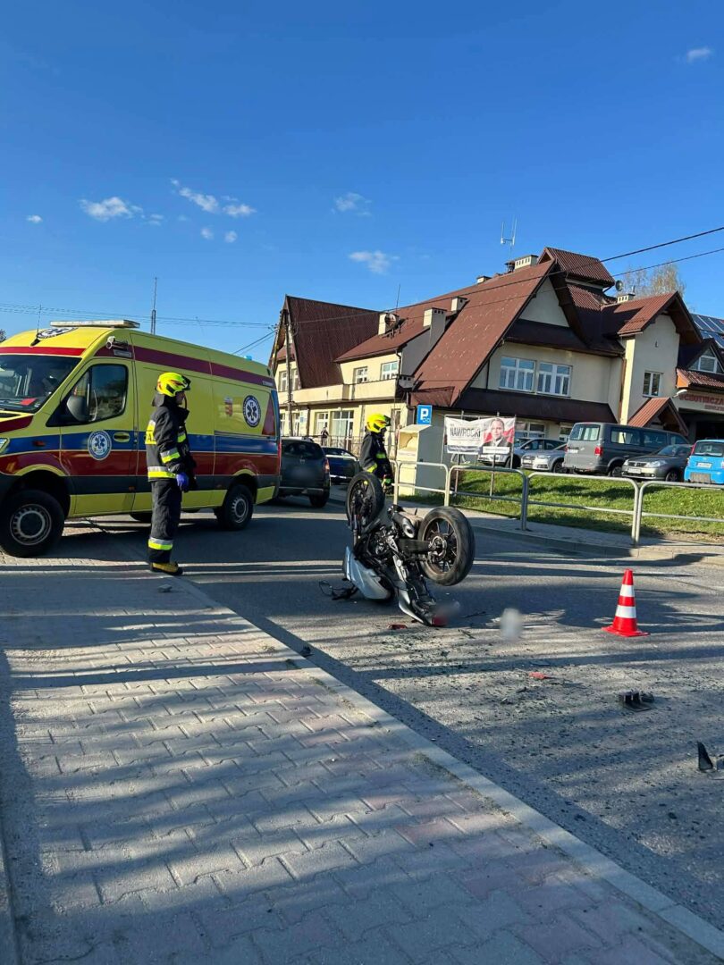Emergency responders and a yellow ambulance at a street crash scene with a toppled motorcycle and traffic cone on a crosswalk.