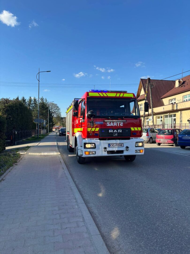 Red fire truck driving down a suburban street on a sunny day, with houses and parked cars along the curb on the right.