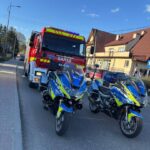 Fire truck and two police motorcycles parked on a sunny street, emergency lights visible in front of a row of buildings