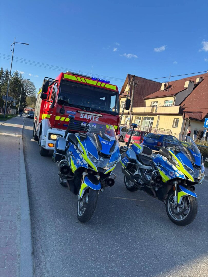 Fire truck and two police motorcycles parked on a sunny street, emergency lights visible in front of a row of buildings