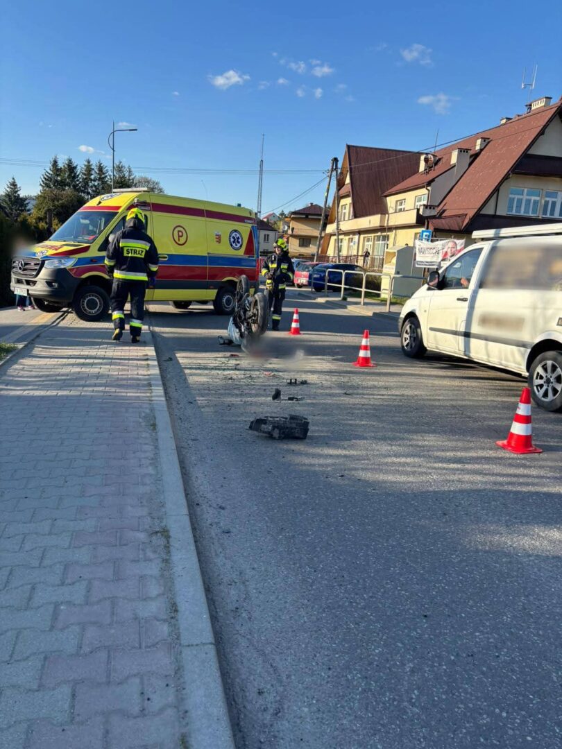 Emergency responders and a yellow ambulance at a street accident, with a toppled motorcycle and traffic cones on the road.