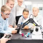 Group of children gathered around a table watching a robotic arm demonstration in a bright lab classroom.