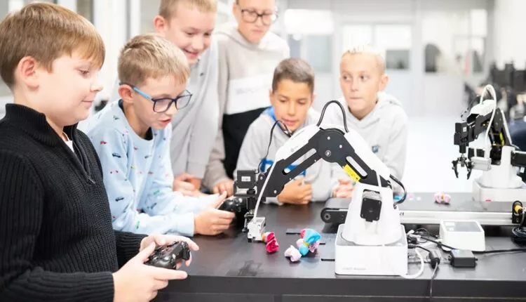 Group of children gathered around a table watching a robotic arm demonstration in a bright lab classroom.