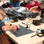Young student wearing safety glasses soldering components at a lab bench with a grid cutting mat and soldering iron in the foreground