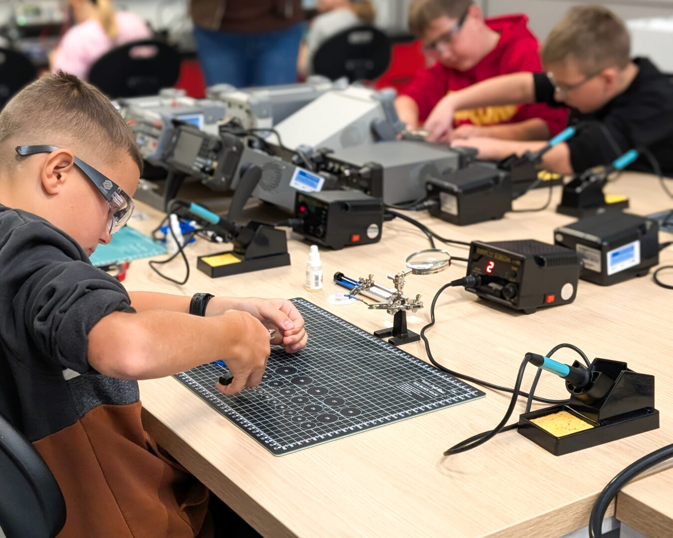 Young student wearing safety glasses soldering components at a lab bench with a grid cutting mat and soldering iron in the foreground