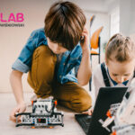 Two kids building a small robot on the floor with a laptop nearby, FABLAB logo visible in the top-left corner of the image, indoors.