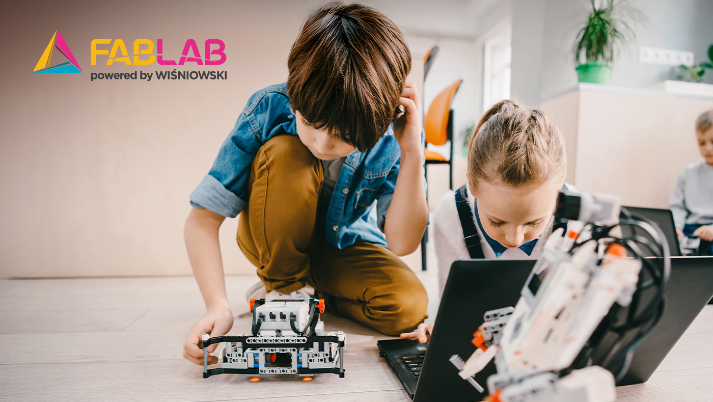 Two kids building a small robot on the floor with a laptop nearby, FABLAB logo visible in the top-left corner of the image, indoors.