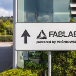 Directional sign: FABLAB powered by Wiśniowski with an upward arrow, outdoors beside a glass building.