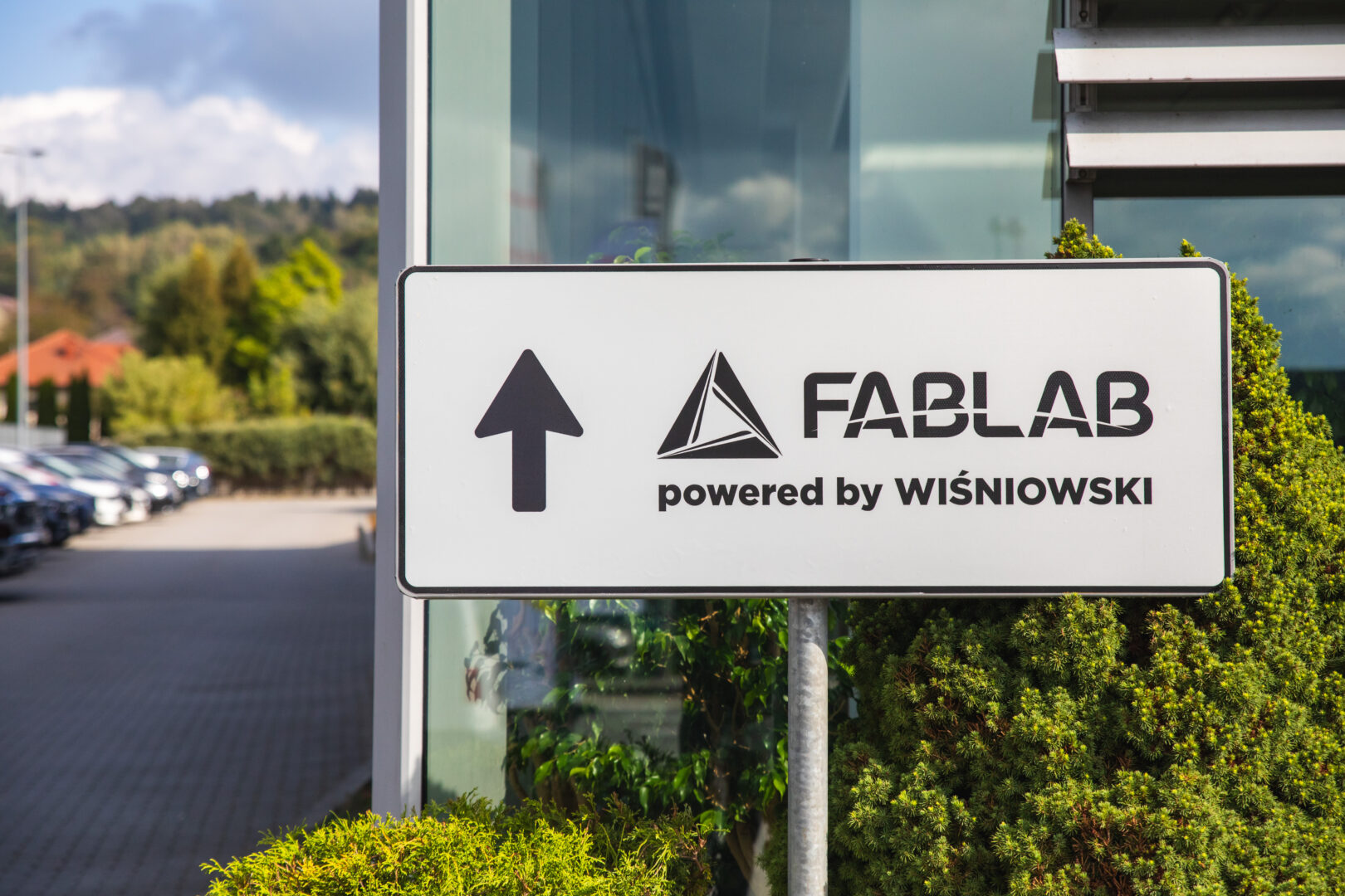 Directional sign: FABLAB powered by Wiśniowski with an upward arrow, outdoors beside a glass building.