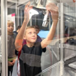 Boy reaching up to touch a robotic arm inside a glass enclosure, surrounded by classmates during a science demonstration