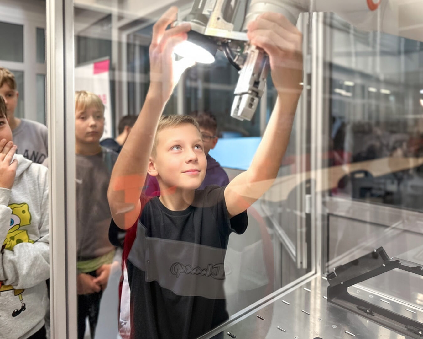 Boy reaching up to touch a robotic arm inside a glass enclosure, surrounded by classmates during a science demonstration