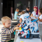 Toddler in a striped shirt builds with a colorful robotics toy at an outdoor table, with adults chatting in the blurred background.