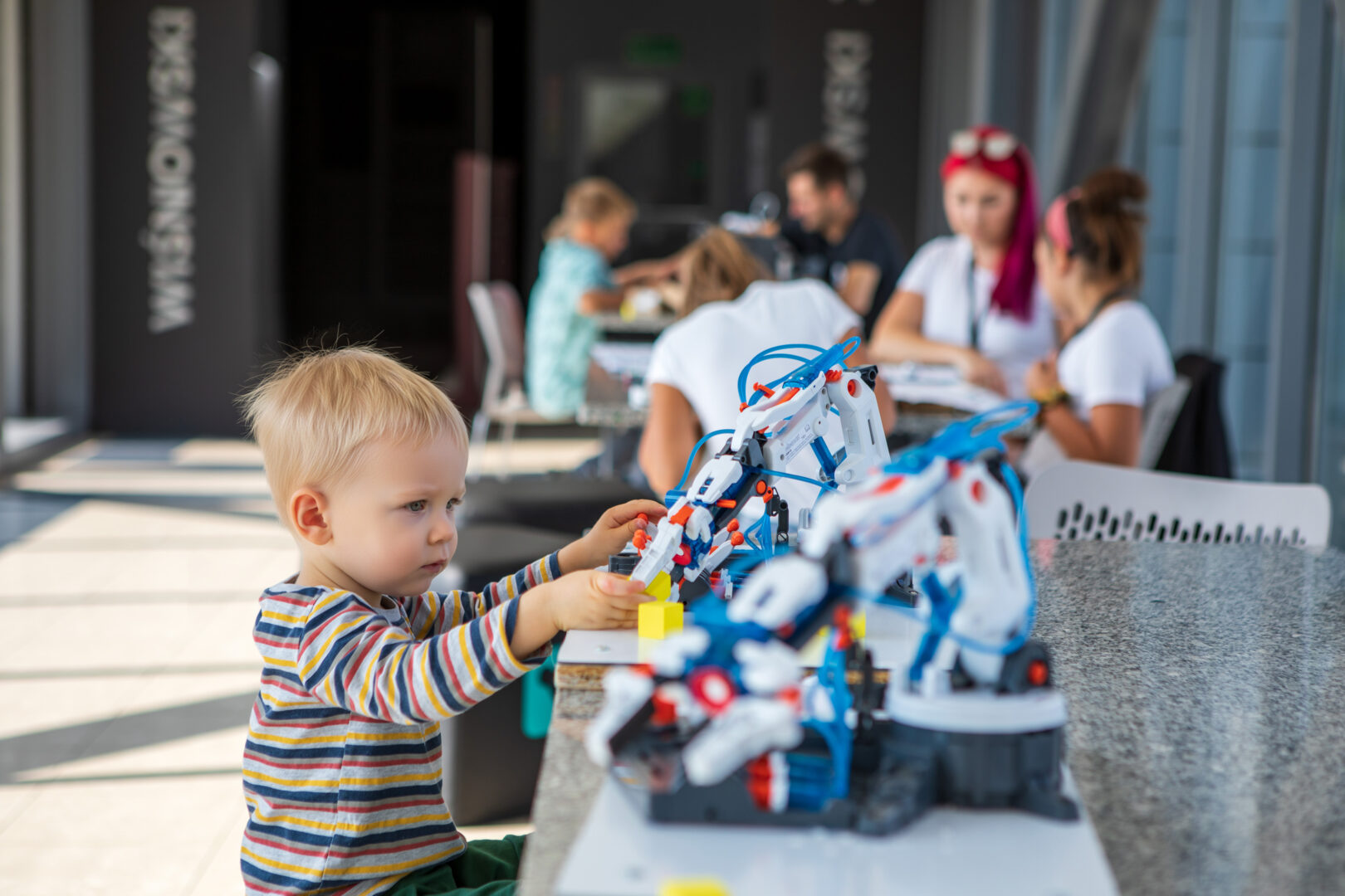 Toddler in a striped shirt builds with a colorful robotics toy at an outdoor table, with adults chatting in the blurred background.