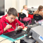 Boy in a red hoodie and safety goggles soldering a circuit on a workbench in a classroom electronics lab, with a magnifier nearby and other students blurred in the background.