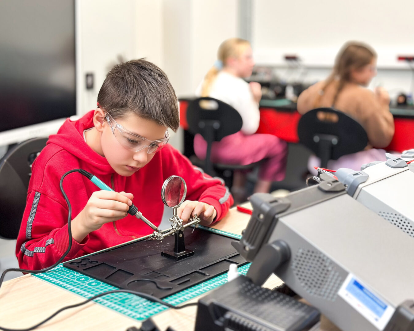 Boy in a red hoodie and safety goggles soldering a circuit on a workbench in a classroom electronics lab, with a magnifier nearby and other students blurred in the background.