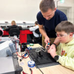Adult mentor helps a boy soldering electronics at a workstation in a makerspace.