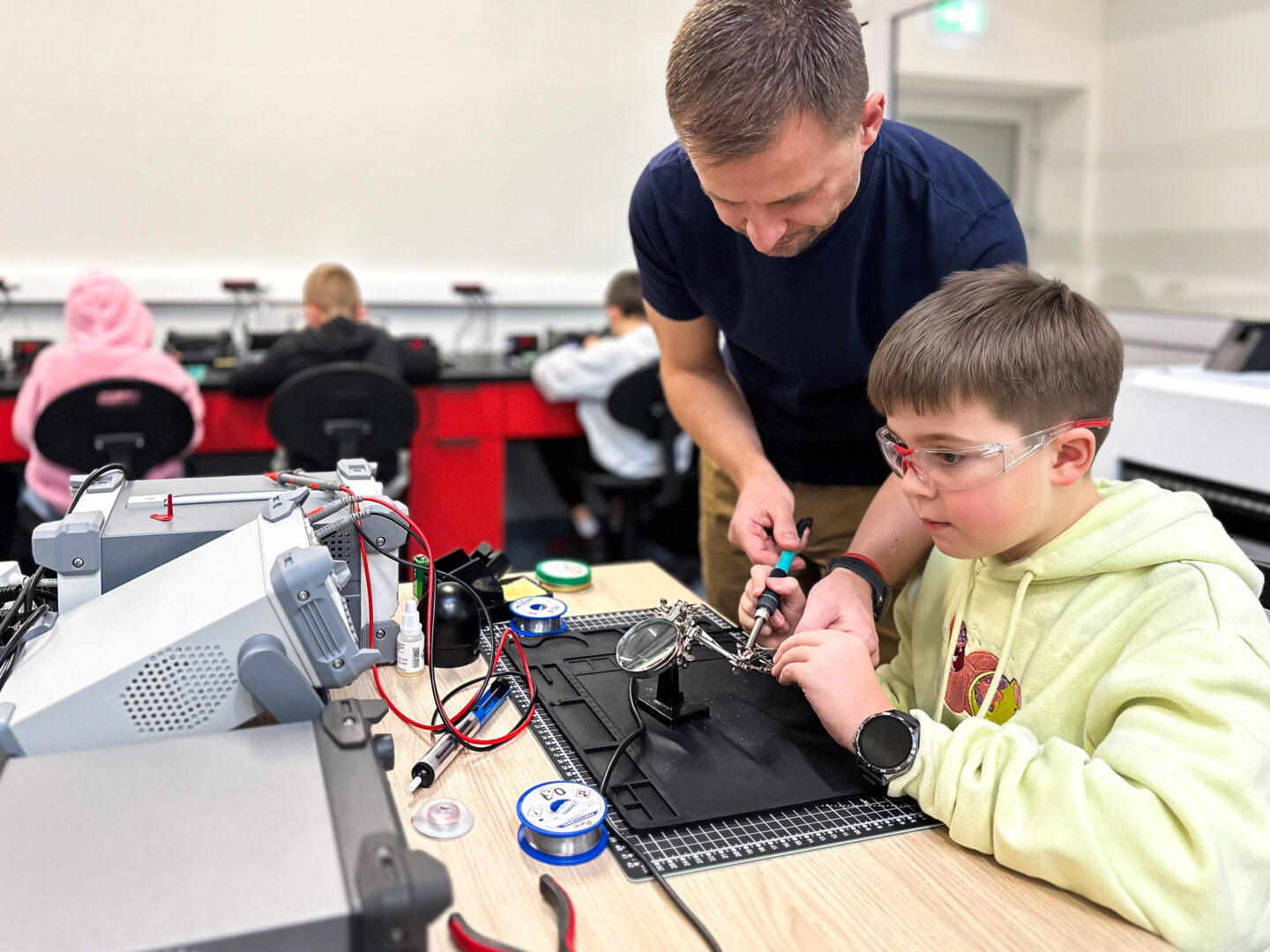 Adult mentor helps a boy soldering electronics at a workstation in a makerspace.