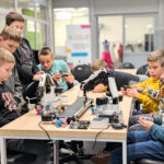Group of boys at a workshop table working with robotic arms and small colorful figures.