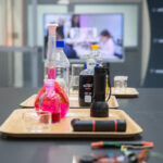 Lab bench with colorful liquids in glass bottles and scientific equipment on a tray, blurred monitor in background.
