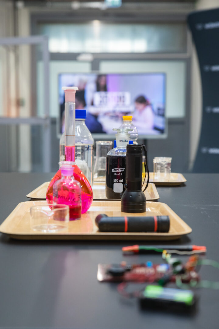Lab bench with colorful liquids in glass bottles and scientific equipment on a tray, blurred monitor in background.