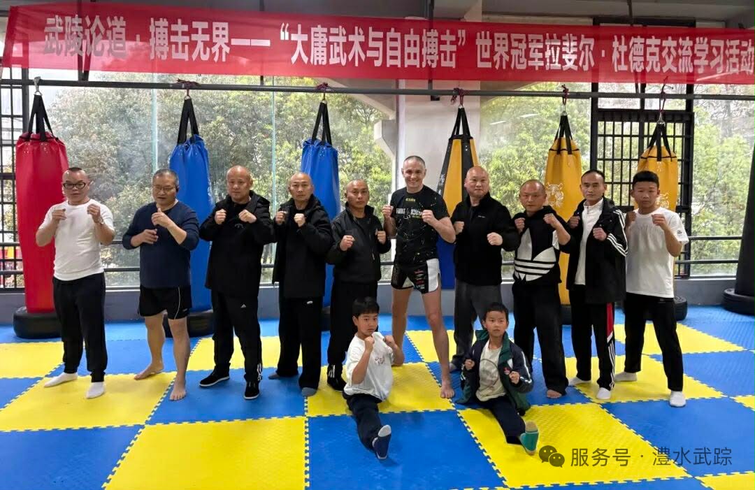 Group of men (and two kids) posing in a martial arts gym with punching bags and a red banner overhead in Chinese.