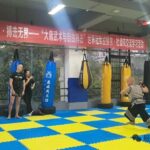 Man practicing a kick in a martial arts gym with blue/yellow padded floor and hanging punching bags; several onlookers watch nearby show a banner in Chinese above the area.