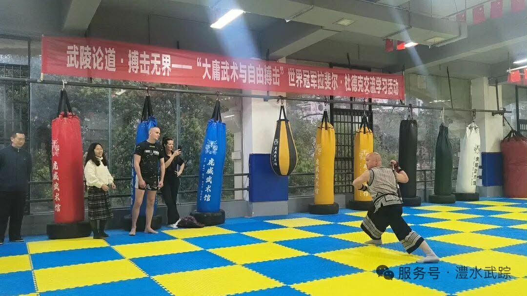 Man practicing a kick in a martial arts gym with blue/yellow padded floor and hanging punching bags; several onlookers watch nearby show a banner in Chinese above the area.