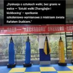 Indoor martial arts training space with red, blue, and yellow punching bags and a red banner with white Chinese characters hanging across the back wall. A text overlay in Polish describes a discussion about martial arts.