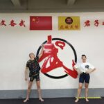 Two people in a martial arts gym pose in front of a large red and black circular emblem on the wall, with Chinese flags and banners above.